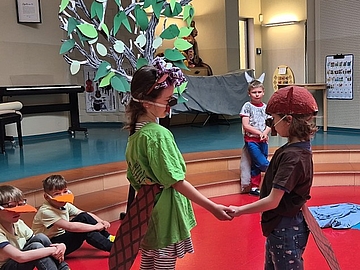Zwei Kinder in selbst gestalteten Kostümen spielen eine Szene aus dem Theaterstück „Kleiner Biber – du schaffst das!“ auf einer Bühne in der Kita. Im Hintergrund sind weitere Kinder sowie ein großer gebastelter Baum als Kulisse zu sehen.